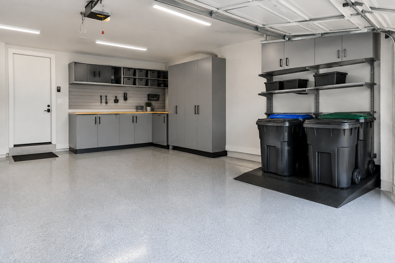 Four wheeled trash cans on a ramped black platform in a corner garage staging area near the driveway